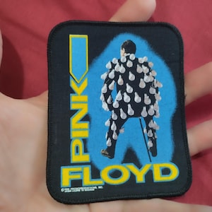 Puede incluir: Un parche rectangular de tela negra con el gráfico de una persona en traje cubierta de bombillas. El texto PINK FLOYD está impreso en amarillo con un borde azul. El parche tiene un borde cosido en negro y un pequeño texto de copyright abajo.