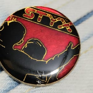 Könnte beinhalten: Eine schwarze und rote Emaille-Anstecknadel mit goldener Umrandung. Die Anstecknadel zeigt eine Silhouette einer Person mit dem Wort "Styx" in goldenen Buchstaben über der Person. Das Wort "Styx" ist von einem roten und goldenen Schlangendesign umgeben.