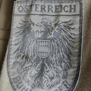 May include: A gray embroidered patch with the text "OSTERREICH" and a coat of arms featuring an eagle with a shield. The text "BUNDESHEER" is visible below the eagle.