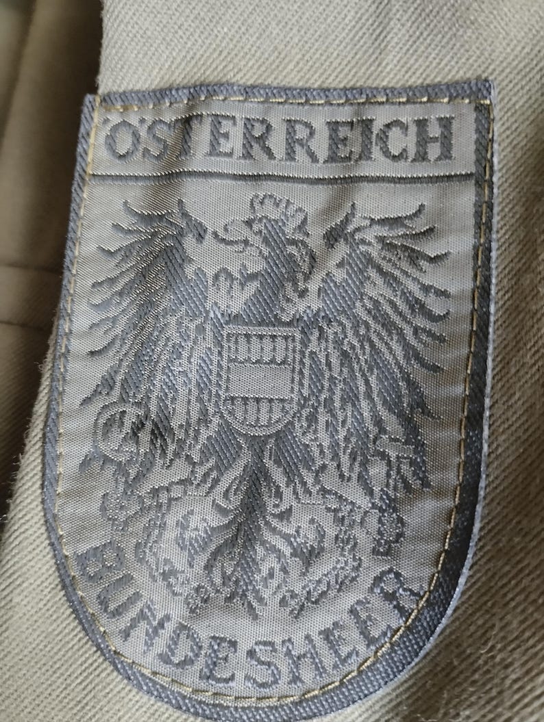May include: A gray embroidered patch with the text "OSTERREICH" and a coat of arms featuring an eagle with a shield. The text "BUNDESHEER" is visible below the eagle.