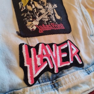 Könnte beinhalten: Nahaufnahme einer hell gewaschenen Jeansjacke mit zwei Aufnähern. Ein Aufnäher zeigt eine Bandillustration mit dem Text "Judas Priest". Der andere Aufnäher hat das Wort "SLAYER" in rosa und weißer Schrift.