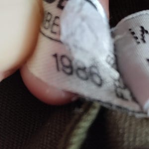 May include: Close-up of a white tag with black text that reads "1986". The tag is attached to a brown and green garment.