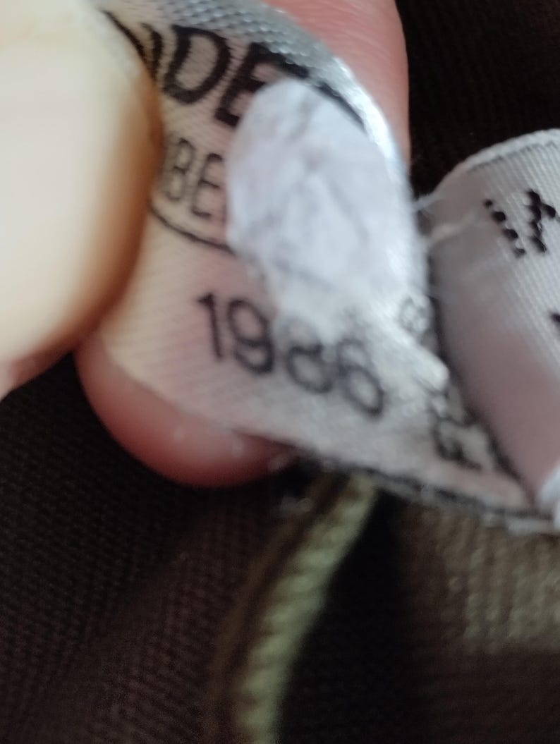 May include: Close-up of a white tag with black text that reads "1986". The tag is attached to a brown and green garment.