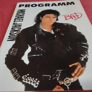 May include: A Michael Jackson Bad tour programme booklet featuring a cover image of the artist wearing a black jacket with multiple silver buckles and zippers. The text reads PROGRAMM, MICHAEL JACKSON, and BAD in red script on a white background.