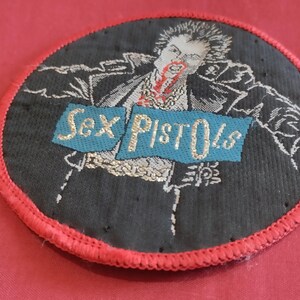 May include: Embroidered patch featuring a black and white illustration of a figure with red accents, with the text "Sex Pistols" on a blue banner. The patch has a red stitched border and is circular in shape.