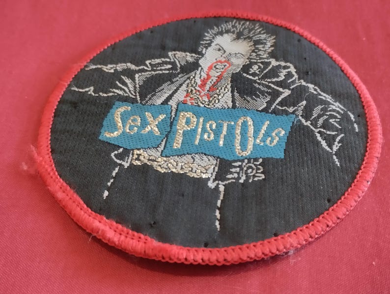 May include: Embroidered patch featuring a black and white illustration of a figure with red accents, with the text "Sex Pistols" on a blue banner. The patch has a red stitched border and is circular in shape.