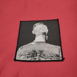 Peut inclure: Écusson noir et blanc représentant une personne avec le logo AC/DC sur la tête. Du texte est visible sur le bord inférieur. L'écusson est posé sur un fond rouge.
