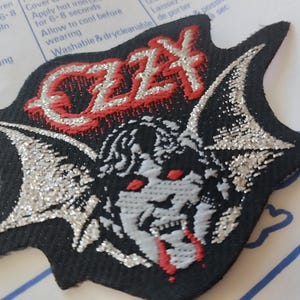 Könnte beinhalten: Schwarzer, sternförmiger Aufnäher mit dem Wort "OZZY" in Rot und Silber. Er zeigt eine detaillierte Illustration eines dämonischen Gesichts mit roten Augen und silbernen Flügeln. Der Aufnäher ist zum Aufbügeln auf Kleidung gedacht.