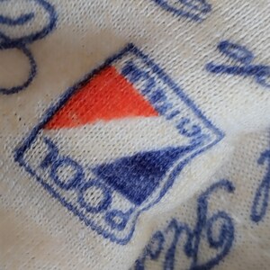 May include: Close-up of a white knitted fabric with a blue and red embroidered logo that says "ROCO" in a blue cursive font.
