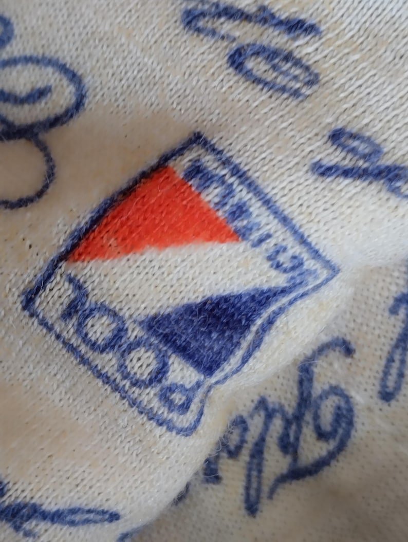 May include: Close-up of a white knitted fabric with a blue and red embroidered logo that says "ROCO" in a blue cursive font.
