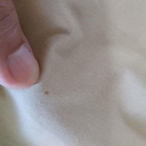 Puede incluir: Primer plano de una tela beige claro con sutiles l&iacute;neas horizontales. Una peque&ntilde;a mancha marr&oacute;n oscuro es visible en la superficie de la tela. La imagen tambi&eacute;n muestra una parte de un dedo.