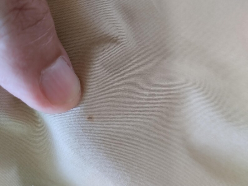 Puede incluir: Primer plano de una tela beige claro con sutiles l&iacute;neas horizontales. Una peque&ntilde;a mancha marr&oacute;n oscuro es visible en la superficie de la tela. La imagen tambi&eacute;n muestra una parte de un dedo.
