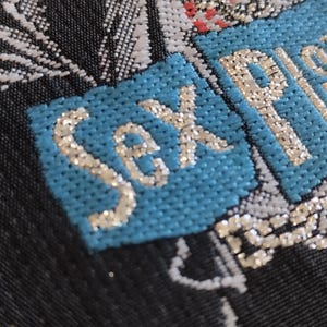 May include: Close-up of a textile with the words "Sex Pistols" embroidered in silver thread on a light blue patch. The background is a dark fabric with white and silver floral patterns.