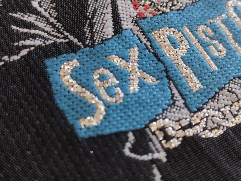 May include: Close-up of a textile with the words "Sex Pistols" embroidered in silver thread on a light blue patch. The background is a dark fabric with white and silver floral patterns.