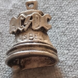 Può includere: Un ciondolo in metallo color argento a forma di campana. La parte superiore presenta la scritta AC/DC in rilievo, mentre la sezione inferiore include il testo HELLS BELL. Il ciondolo ha un piccolo occhiello in cima e un battaglio arrotondato.