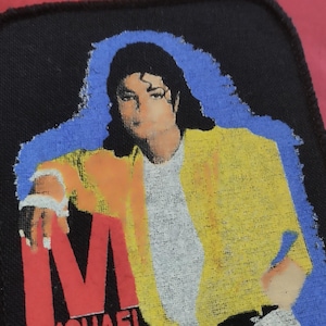 May include: A rectangular fabric patch with a black border featuring a graphic print of Michael Jackson. The figure wears a yellow jacket over a white shirt, set against a blue background. The red letter M is visible at the bottom left corner.