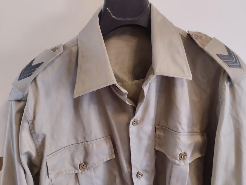 May include: A beige long-sleeved shirt with black chevron patches on the shoulders and a button-up front.