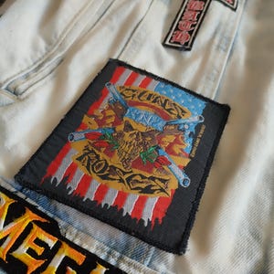 May include: Pale blue denim jacket with patches. One patch displays the Guns N' Roses logo with crossed guns and a skull over an American flag design. Other patches include a cross and the word "MEGADETH" in orange and yellow.