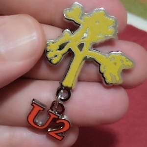 Op de afbeelding: Een metalen speld in de vorm van een gele Joshua-boom met een zilverkleurige omlijning. Een kleinere, losse zilverkleurige bedel met de oranje tekst U2 is met een klein metalen ringetje aan de onderkant van de boom bevestigd voor een hangend effect.