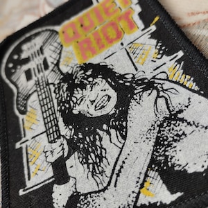 Peut inclure: Un écusson en tissu noir présentant une illustration graphique blanche d'une personne aux cheveux longs tenant une guitare basse. Le texte QUIET RIOT est imprimé en jaune en haut. Le motif inclut des accents hachurés jaunes sur fond noir.