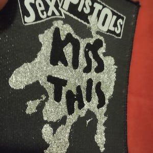Może przedstawiać: Czarna naszywka z napisami "Sex Pistols" i "Kiss This" w białych i czarnych literach. Napisy znajdują się na tle srebrnego brokatu. Naszywka jest prostokątna.