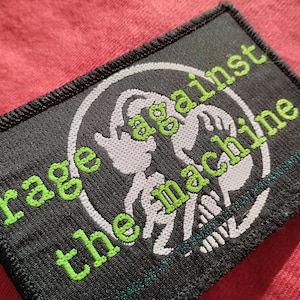 Może przedstawiać: Czarna prostokątna naszywka z napisem "rage against the machine" w kolorze zielonym, z białym graficznym przedstawieniem szkieletu. Naszywka znajduje się na czerwonej powierzchni. Naszywka ma czarną obwódkę.
