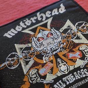 Pode incluir: Patch quadrado preto com o nome da banda "Motörhead" em branco, acima de uma ilustração detalhada. A ilustração apresenta uma caveira com olhos vermelhos, asas e uma cruz. O texto "ALL THE ACES" está abaixo.