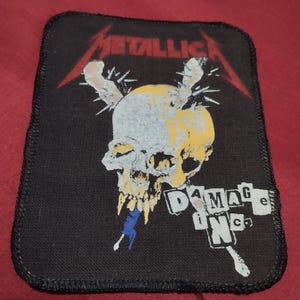 Op de afbeelding: Zwarte patch met het Metallica-logo in rood en een schedelafbeelding met gele en blauwe accenten. De tekst "DAMAGE INC" is in het wit gedrukt. De patch heeft een gestikte rand.