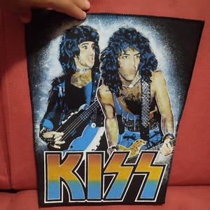 Könnte beinhalten: Ein rechteckiger Stoffaufnäher mit einem Grafikdruck von zwei Rockmusikern mit langem dunklem Haar, von denen einer einen blauen Bass und der andere eine Gitarre spielt. Unter den Figuren ist das Wort KISS in einer fetten Schrift gedruckt.