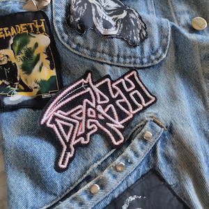 Może przedstawiać: Jasnoniebieska kamizelka dżinsowa z kilkoma haftowanymi naszywkami, w tym prostokątną naszywką Megadeth z żółtym tekstem, czarno-białą naszywką z czaszką oraz czarną naszywką z białym napisem Death. Kamizelka posiada srebrne metalowe guziki.