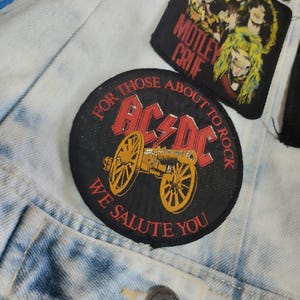 Puede incluir: Chaqueta vaquera azul claro con varios parches. El parche principal es redondo, negro y presenta el texto "AC/DC" en rojo con un diseño de cañón amarillo y las palabras "FOR THOSE ABOUT TO ROCK WE SALUTE YOU".