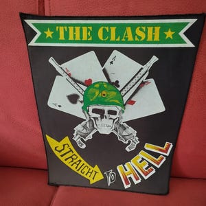 Op de afbeelding: Zwarte patch met een schedel met een groene helm, gekruiste geweren en speelkaarten. De tekst "THE CLASH" staat erboven en "STRAIGHT TO HELL" eronder. Het ontwerp staat op een zwarte achtergrond.