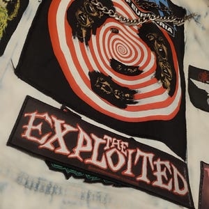 May include: A white denim jacket with various patches and a silver chain. Patches display band logos and graphics, including a black and red spiral design and the words "THE EXPLOITED".