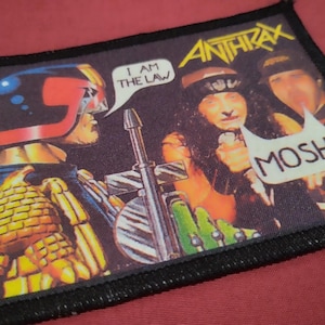 Może przedstawiać: Prostokątna naszywka z tkaniny z grafiką przedstawiającą Judge Dredd i zespół Anthrax. Obraz zawiera dymki z tekstem I AM THE LAW oraz MOSH!, a także nazwę zespołu Anthrax wydrukowaną żółtą czcionką w górnej części naszywki.