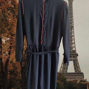 Könnte beinhalten: Ein schwarzes Kleid mit langen Ärmeln und einem passenden Gürtel. Das Kleid hat rote Paspeln am Kragen, der Vorderseite und den Ärmeln. Es hat eine Knopfleiste und ein klassisches Design. Der Eiffelturm ist im Hintergrund zu sehen.