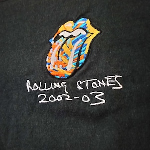 Puede incluir: Camiseta negra con el icónico logo de la lengua y los labios de los Rolling Stones, bordado en colores brillantes. El logo está sobre el texto "Rolling Stones 2002-03" en costuras blancas. La camiseta tiene cuello redondo y cuello gris oscuro.