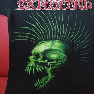 Op de afbeelding: Zwart T-shirt met de rode tekst "THE EXPLOITED". De afbeelding is een groene schedel met een hanenkam en scherpe tanden. Het ontwerp is gecentreerd op de voorkant van het shirt.