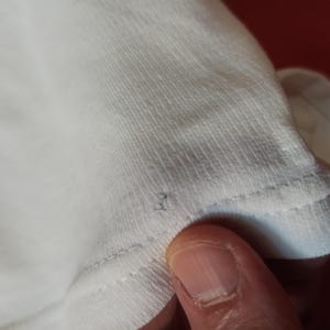 May include: Close-up of white fabric, possibly a t-shirt, with visible stitching along the edge. The fabric appears soft and slightly textured. A finger is visible in the frame, touching the fabric.