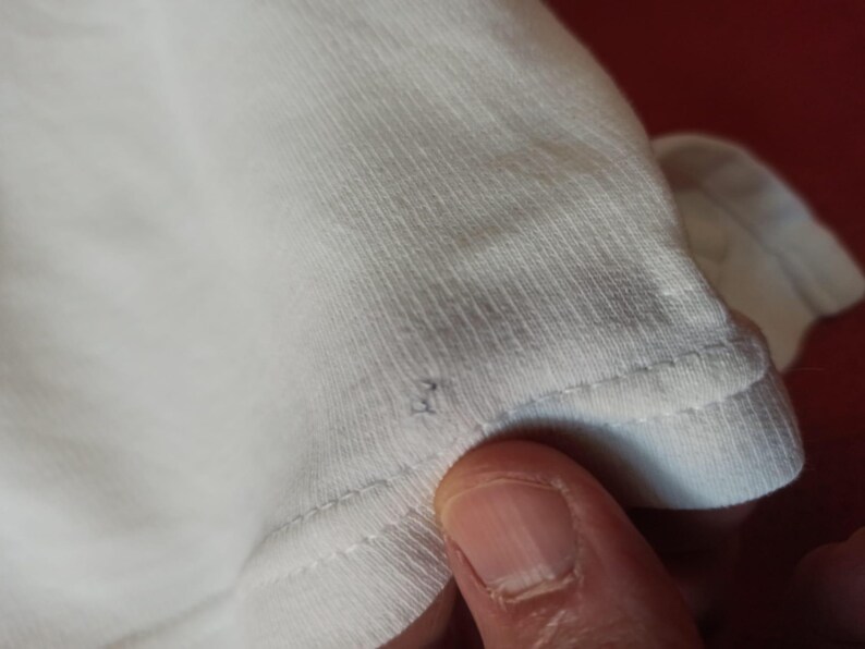 May include: Close-up of white fabric, possibly a t-shirt, with visible stitching along the edge. The fabric appears soft and slightly textured. A finger is visible in the frame, touching the fabric.