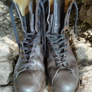 Puede incluir: Un par de botas de combate de cuero marrón oscuro desgastadas con cordones negros. Las botas tienen un diseño de caña alta, costuras visibles y una suela gruesa. El forro interior es de color beige claro. Las botas están sobre una superficie rocosa.