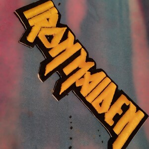 Könnte beinhalten: Gestickter Aufnäher mit dem Text "IRON MAIDEN" in Gelb, umrandet von Schwarz. Der Aufnäher befindet sich auf einem Hintergrund mit blauem, rosa und weißem Batikmuster. Der Aufnäher ist rechteckig mit einem leichten Winkel.