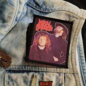 May include: A black and white patch with the text "Metal Church" in red letters. The patch features a photo of two men with long hair, one wearing a black shirt and the other wearing a black shirt and a black hat. The patch is sewn onto a blue denim jacket.