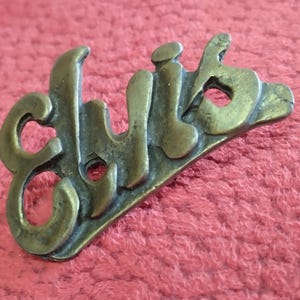 May include: A gold-colored metal pin with the word "Elvis" embossed in a stylized font.