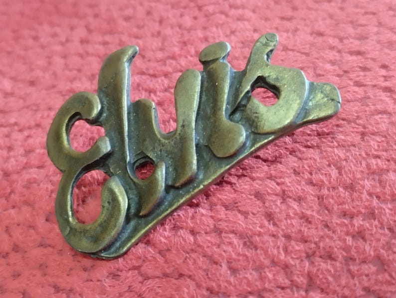 May include: A gold-colored metal pin with the word "Elvis" embossed in a stylized font.
