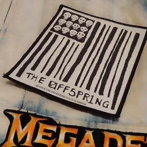 Peut inclure: Gros plan sur une veste en jean avec plusieurs écussons. L'un d'eux présente un drapeau américain noir et blanc avec des crânes. Un autre affiche le nom du groupe "THE OFFSPRING". Un troisième écusson porte le mot "MEGADETH" en orange.