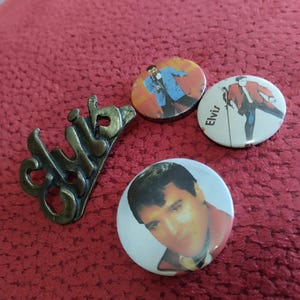May include: Three round pinback buttons featuring images of Elvis Presley. One button has a black and white image of Elvis singing into a microphone. Another button has a color image of Elvis in a blue suit and white shirt. The third button has a color image of Elvis in a red jacket and blue pants. The buttons are on a red fabric surface. There is also a metal pin with the word "club" written on it.
