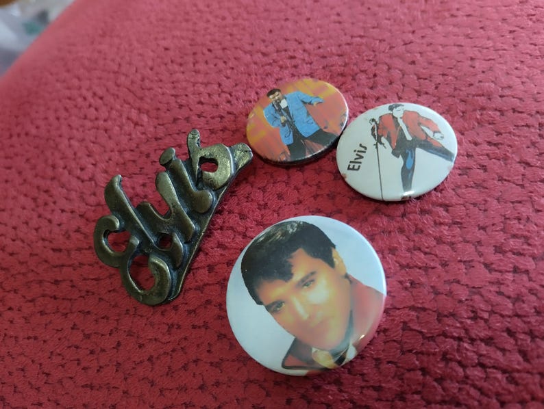 May include: Three round pinback buttons featuring images of Elvis Presley. One button has a black and white image of Elvis singing into a microphone. Another button has a color image of Elvis in a blue suit and white shirt. The third button has a color image of Elvis in a red jacket and blue pants. The buttons are on a red fabric surface. There is also a metal pin with the word "club" written on it.