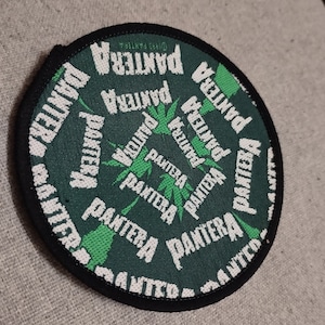 Könnte beinhalten: Ein runder, schwarz umrandeter Patch mit grünem Hintergrund und weißem Text, der in einem Kreislauf immer wieder "PANTERA" lautet. Der Text ist von grünen Palmenblatt-Designs umgeben.