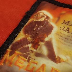 Peut inclure: Un écusson carré avec une bordure noire et une image floue d'une personne en costume. Le mot "MEGADETH" est en lettres orange en bas. Le fond est un mélange d'orange et de marron.