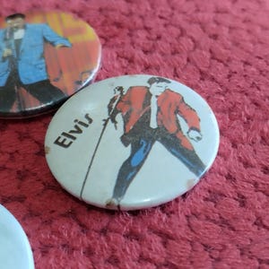 May include: Two round pinback buttons featuring Elvis Presley. The first button has a red, white, and blue background with a black and white image of Elvis Presley in a blue suit. The second button has a white background with a black and white image of Elvis Presley in a red suit. The text "Elvis" is printed in black on the second button.